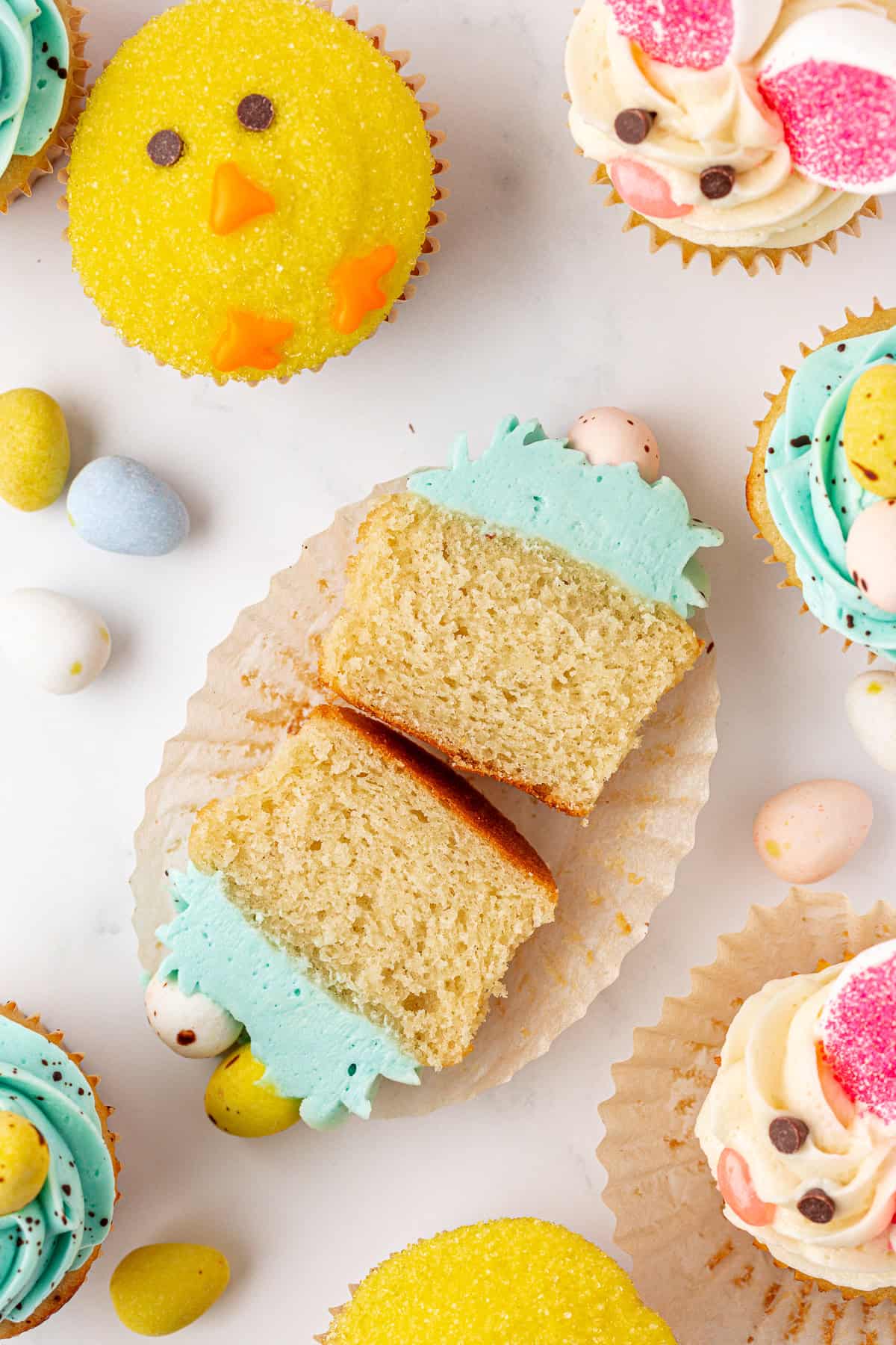 A cupcake with light blue frosting, pastel mini eggs, and a decorative grass design is cut in half on a white surface, surrounded by chick-themed and pastel-decorated cupcakes and candy eggs.