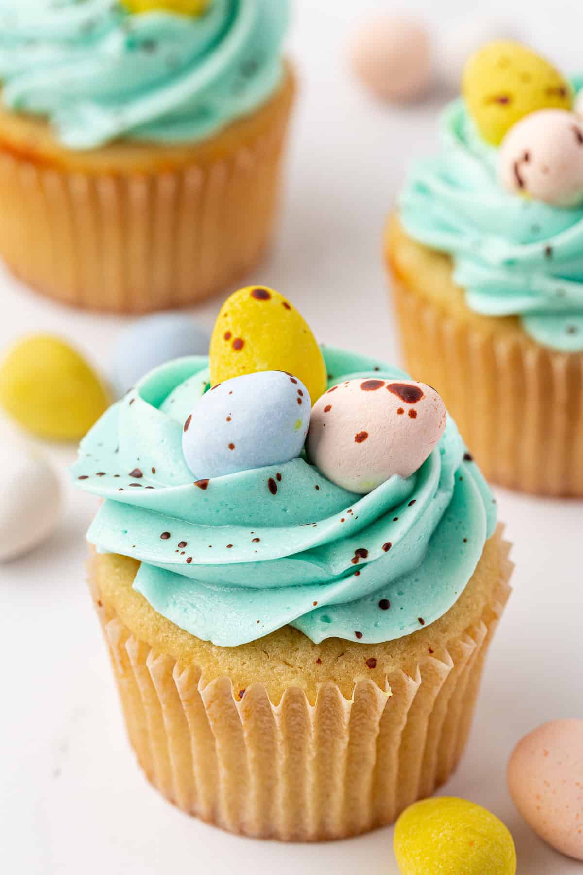A vanilla cupcake with light blue frosting, topped with three pastel-colored candy eggs, sits on a white surface. Other decorated cupcakes and candy eggs are visible in the background.
