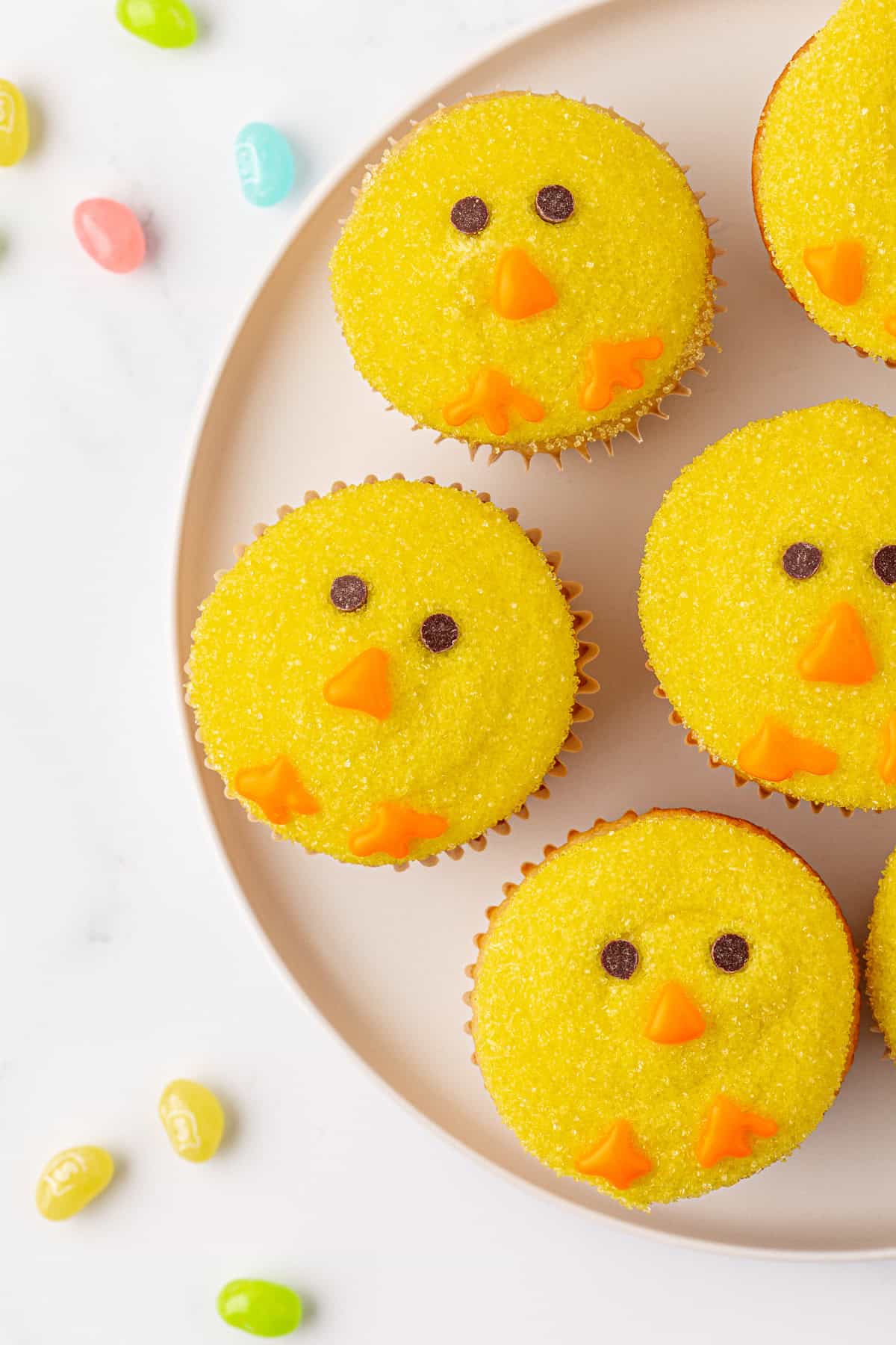 Cupcakes decorated to look like yellow chicks with orange candy beaks and feet, and chocolate eyes, arranged on a round plate. Colorful jelly beans are scattered on the white surface around the plate.