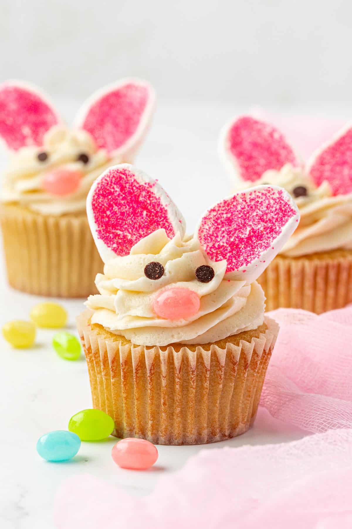 Three cupcakes decorated as bunnies with pink and white candy ears, chocolate chip eyes, and a pink candy nose, surrounded by colorful jellybeans on a white surface with a pink cloth.