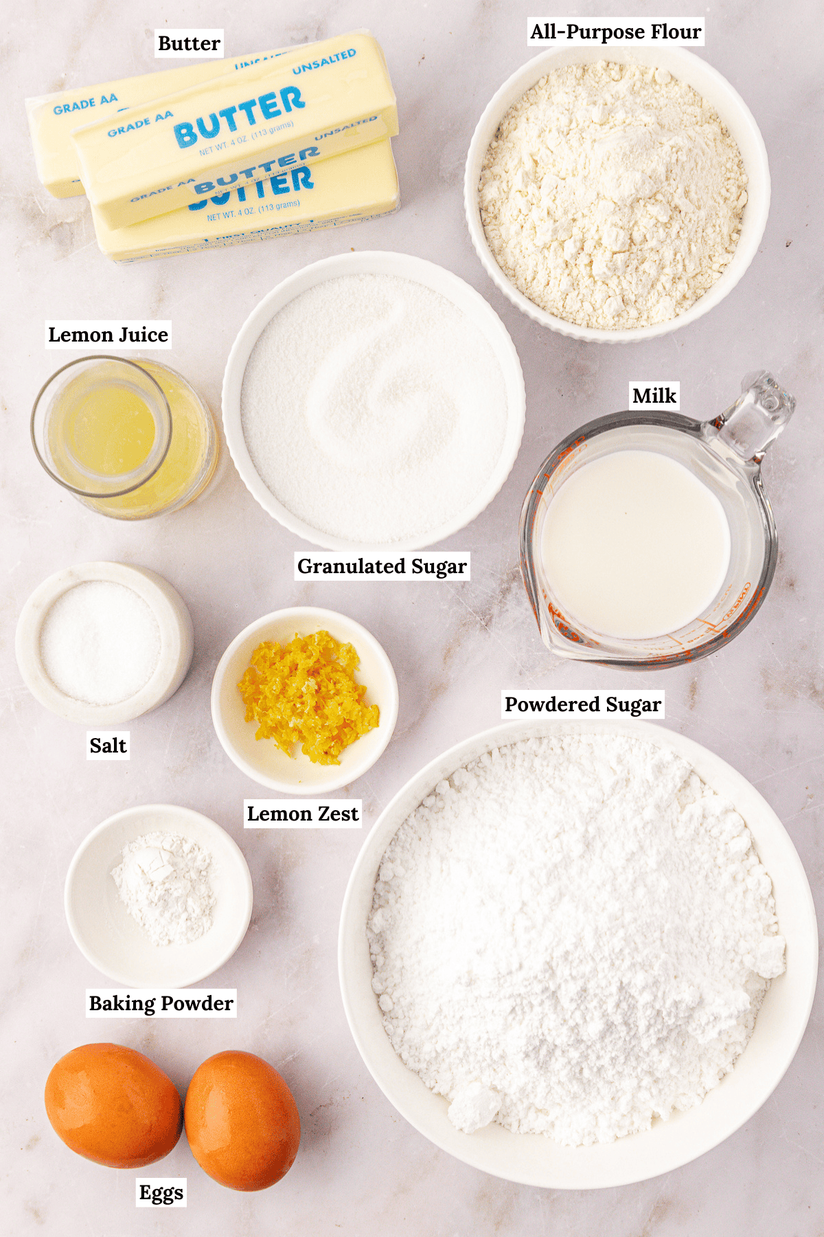 Top-down view of baking ingredients including butter, all-purpose flour, granulated sugar, milk, powdered sugar, eggs, baking powder, salt, lemon zest, and lemon juice, each labeled with text.