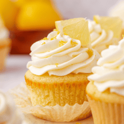 A lemon cupcake topped with creamy white frosting and a lemon slice sits on a marble surface, with other cupcakes and whole lemons blurred in the background. The text Lemon Cupcakes appears at the top.