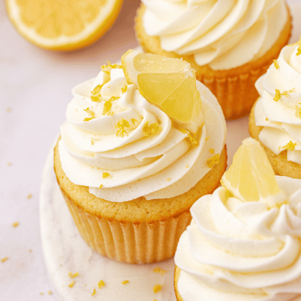 Cupcakes with swirled white frosting are topped with lemon wedges and lemon zest, arranged on a white surface, with a halved lemon visible in the background.