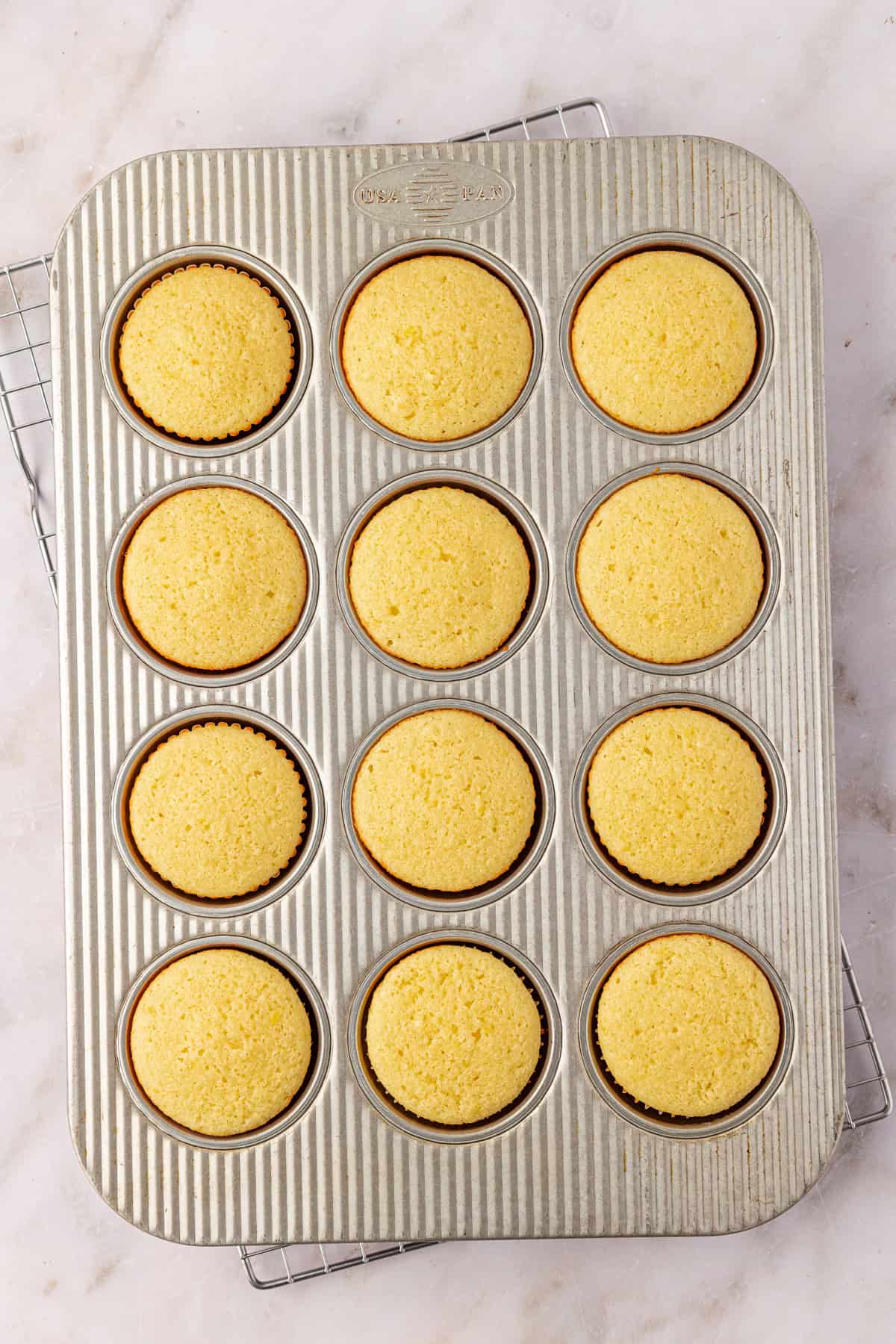 A metal muffin tin with twelve evenly baked yellow cupcakes in paper liners sits on a cooling rack atop a light-colored marble surface.