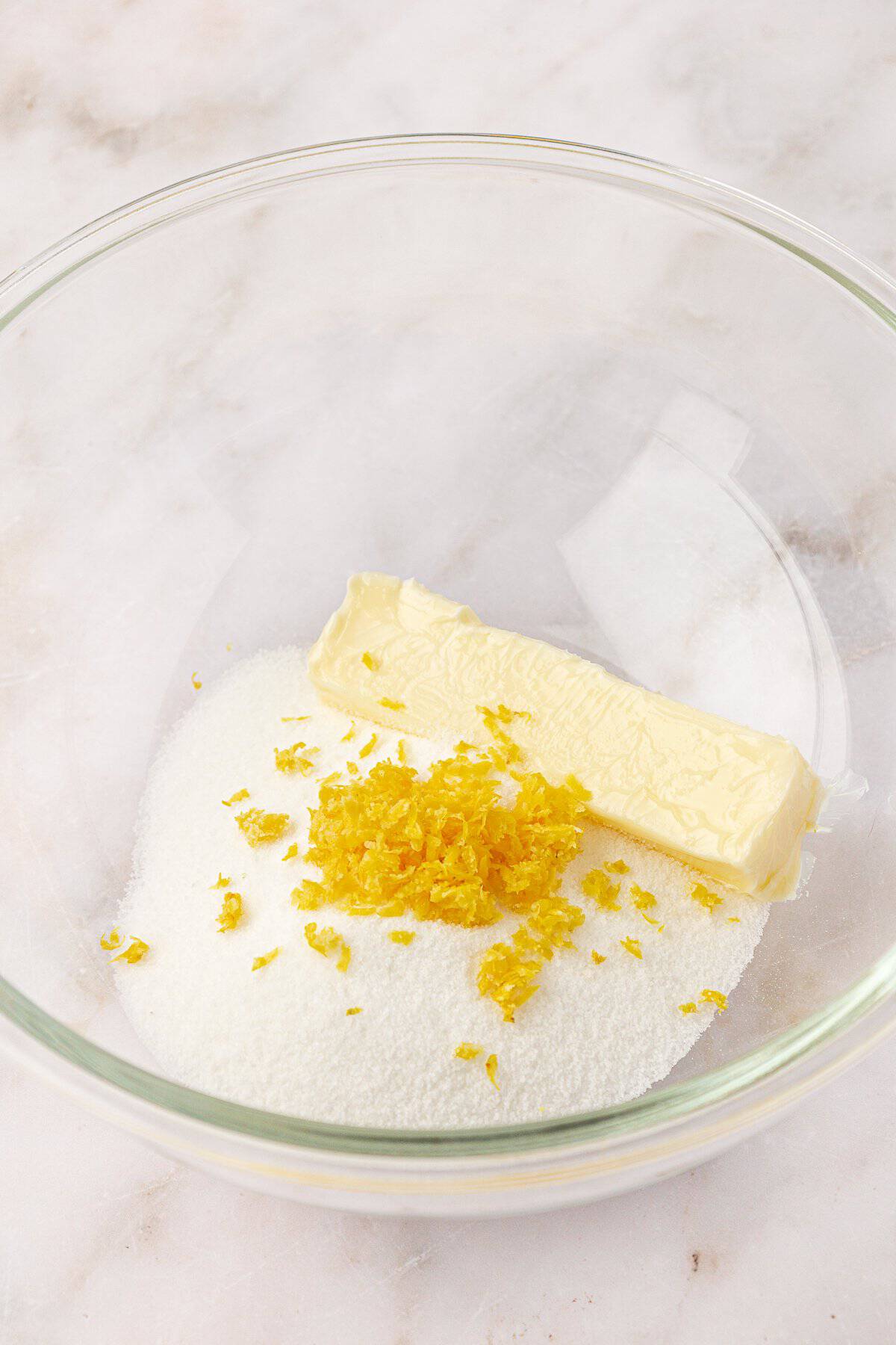 A clear glass bowl containing a stick of butter, granulated sugar, and fresh lemon zest on a marble surface.