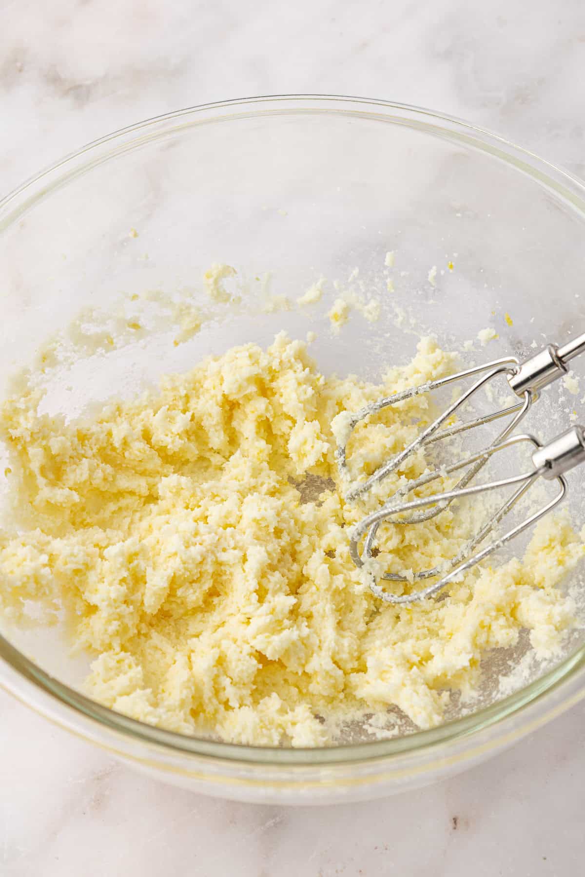 A glass bowl containing a creamed mixture of butter and sugar with a hand mixer resting inside, on a light marble surface.