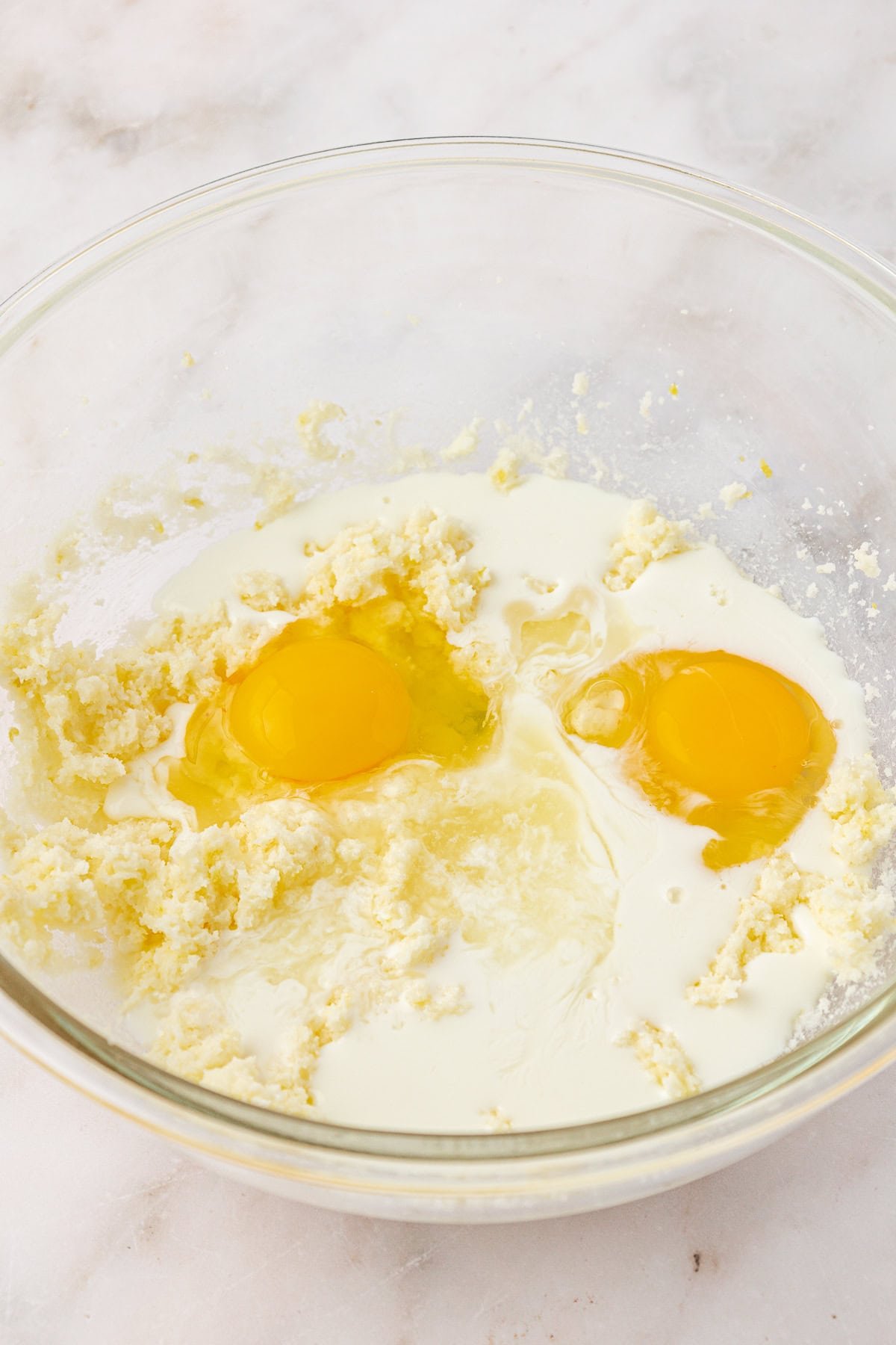 A clear glass bowl contains a mixture of creamed butter and sugar, with two raw eggs and a splash of milk added on top, ready to be mixed together on a light marble surface.