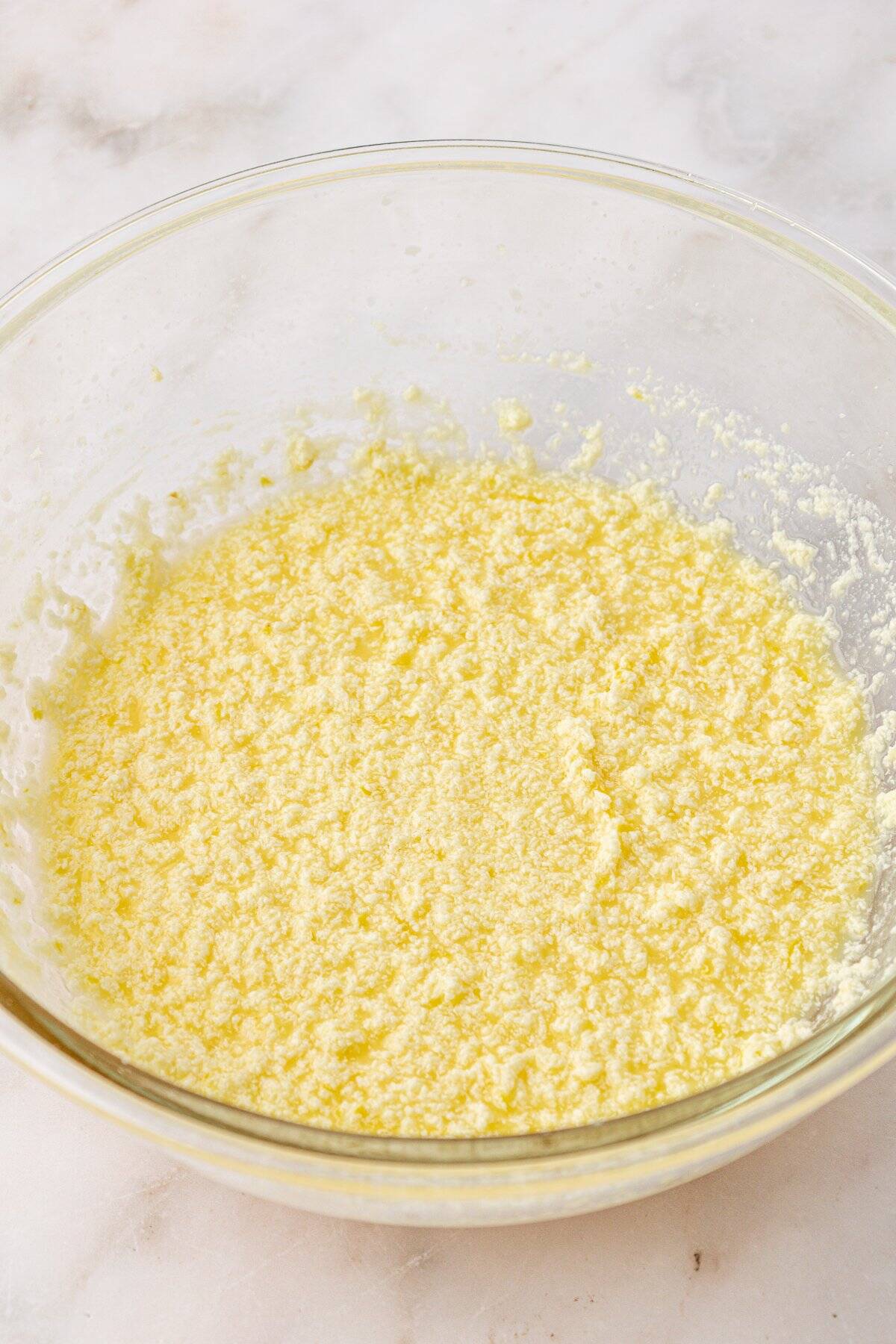 A glass bowl filled with a yellow, grainy mixture sits on a marble surface. The mixture appears to be creamed butter and sugar, often used as a base for baking recipes.
