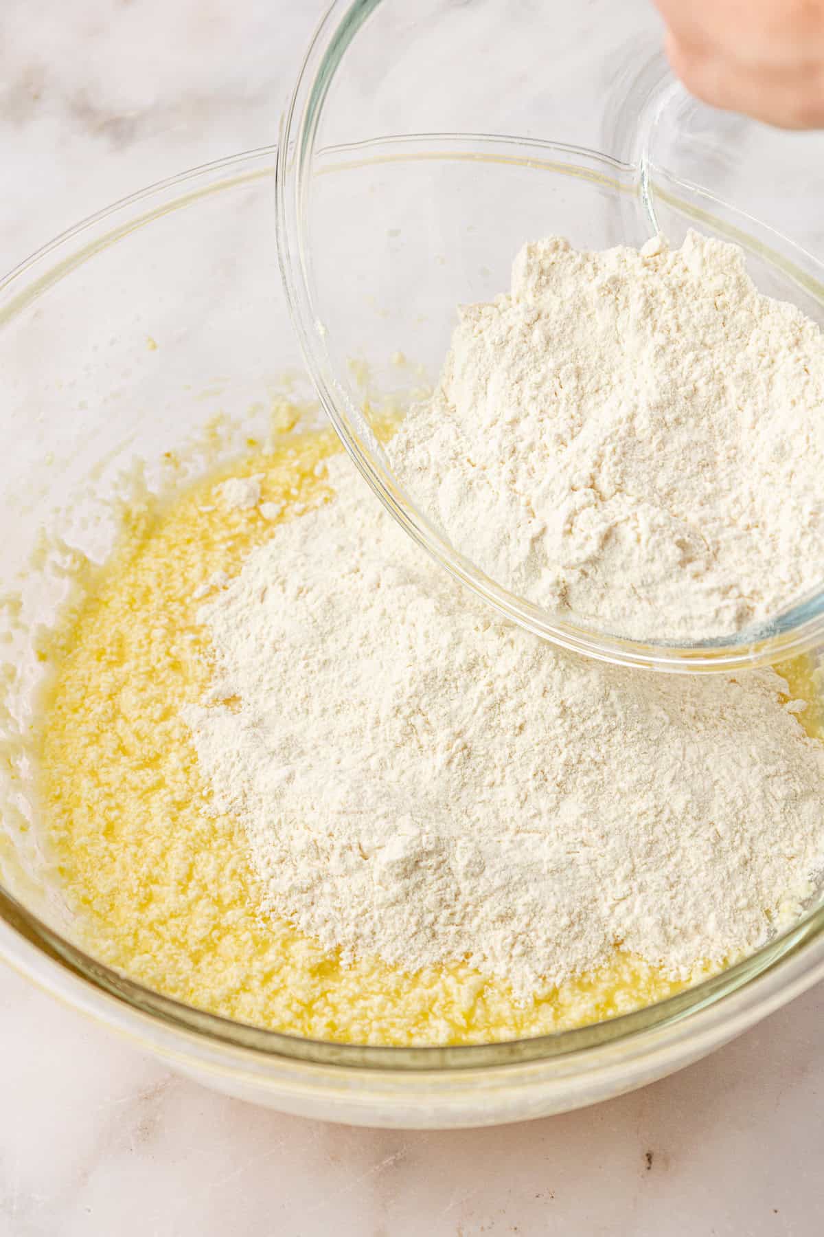 A hand pours flour from a glass bowl into a larger bowl containing a yellow, wet mixture, preparing ingredients for baking on a light-colored surface.