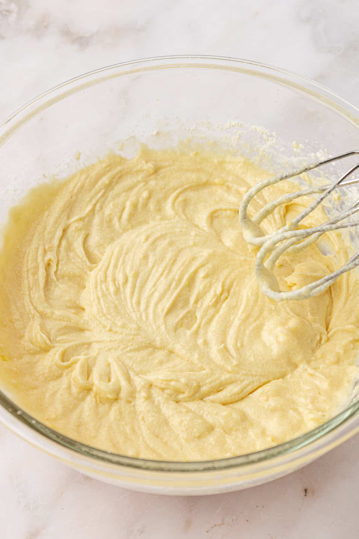 A glass mixing bowl filled with creamy yellow cake batter sits on a marble surface. An electric hand mixer with batter on the beaters rests on the edge of the bowl.