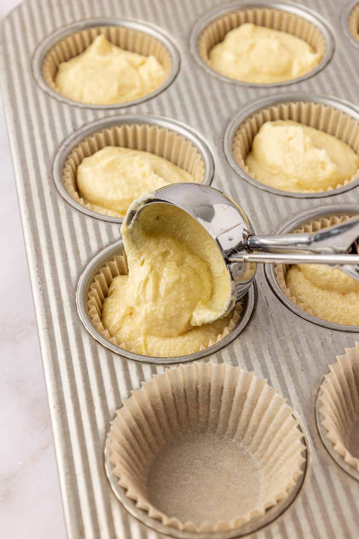 A metal ice cream scoop fills paper cupcake liners in a muffin tin with yellow batter; some liners are filled while others are still empty.