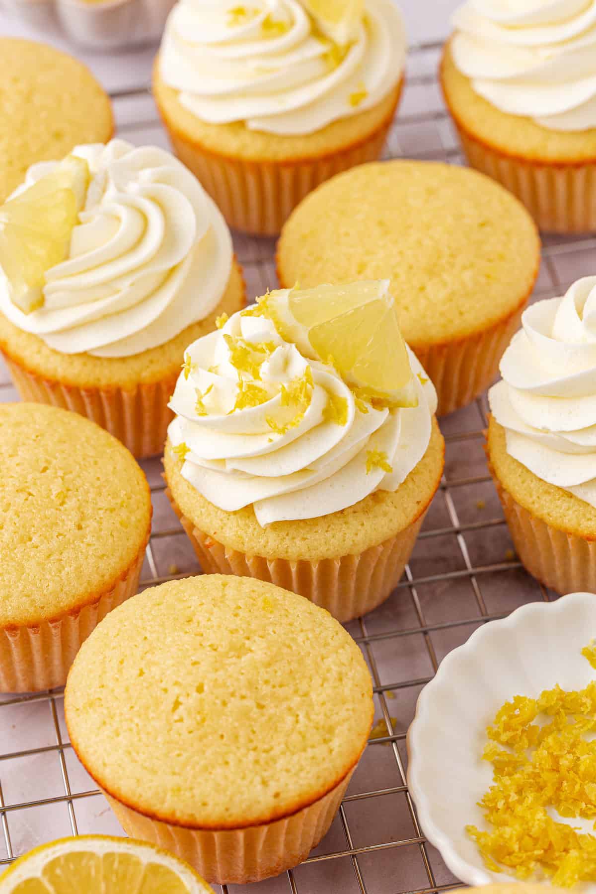 Lemon cupcakes topped with swirls of white frosting, lemon zest, and lemon wedges are arranged on a cooling rack. Some cupcakes are unfrosted. A small dish of lemon zest is nearby.