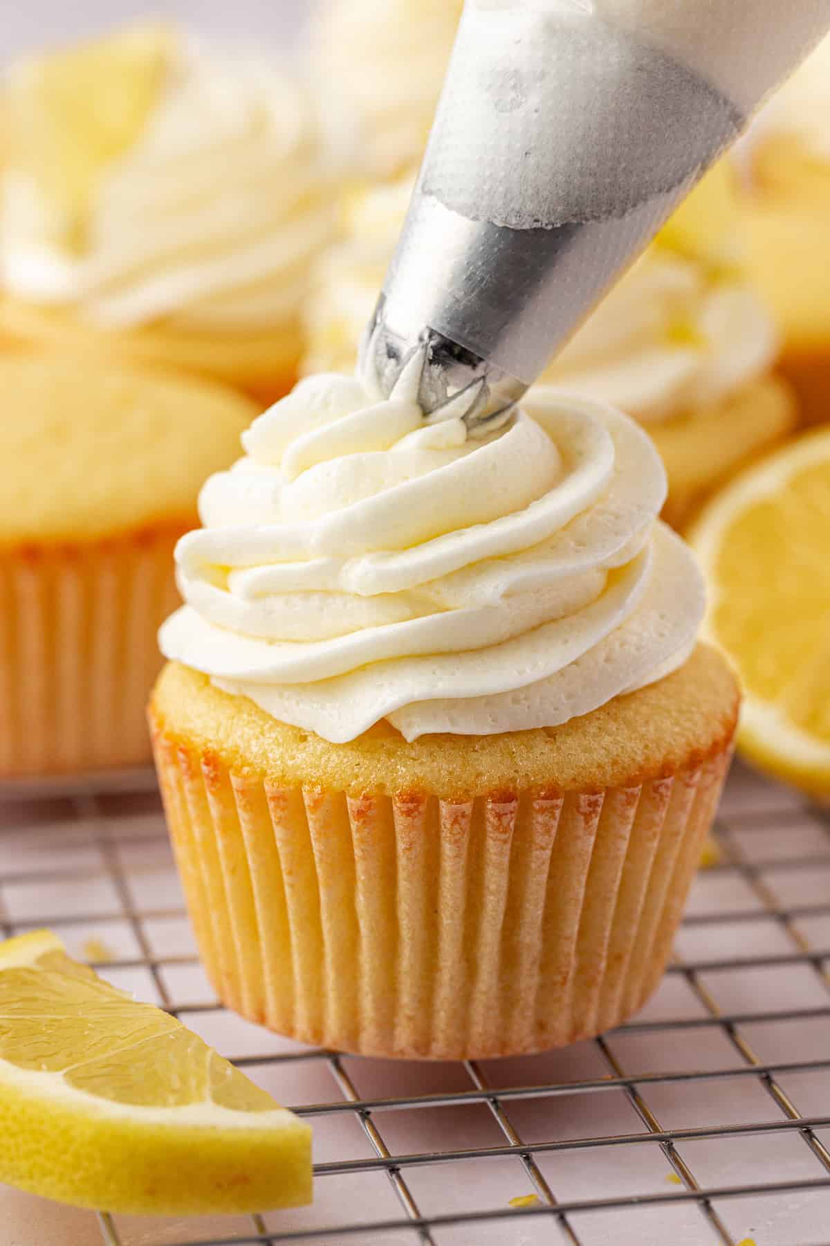 A close-up of a lemon cupcake on a cooling rack being topped with swirls of white frosting from a piping bag, with lemon slices in the background.