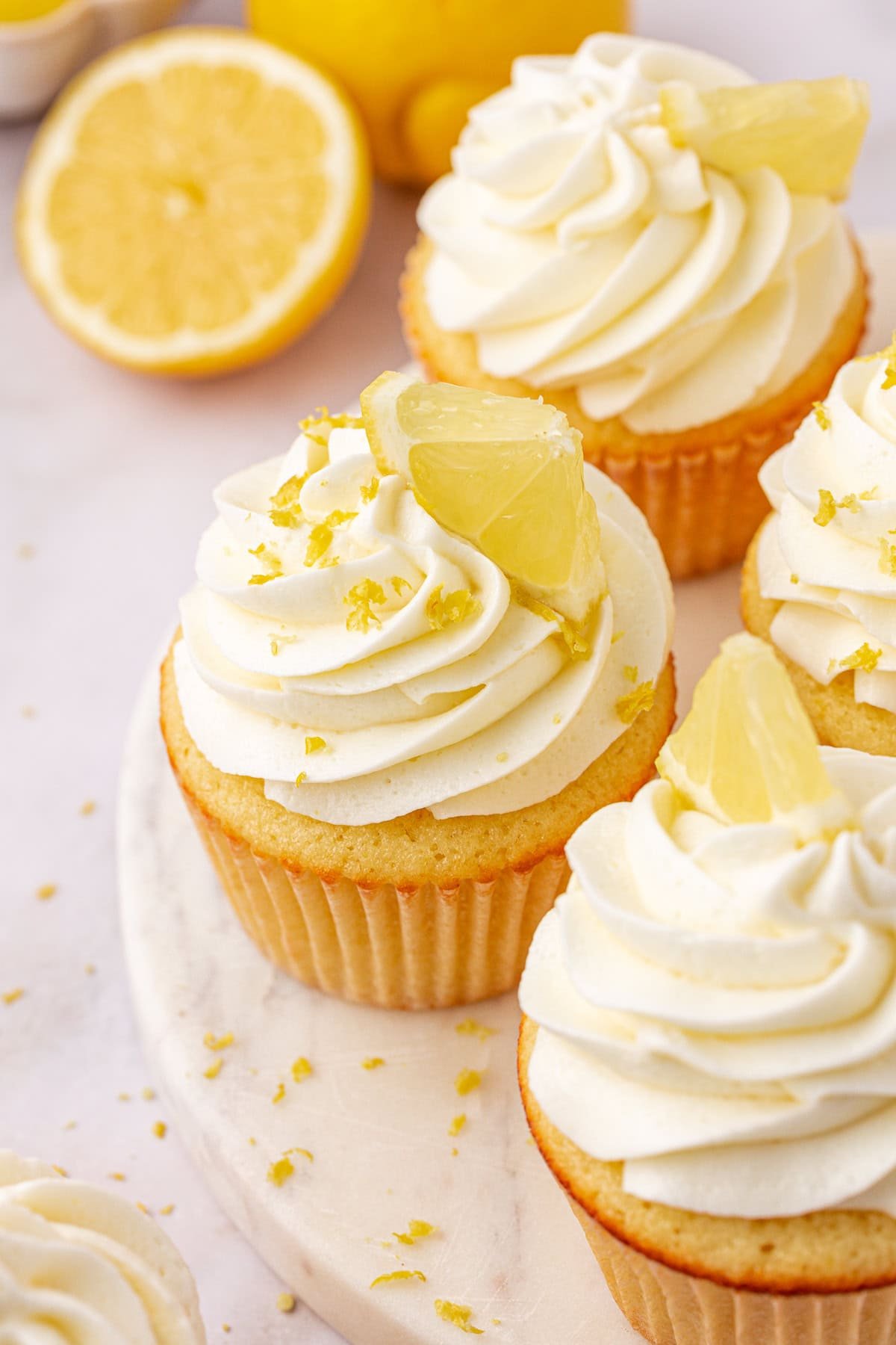 Four lemon cupcakes with swirled white frosting, topped with lemon slices and zest, are arranged on a white surface. In the background, there are halved lemons and additional lemon zest scattered around.