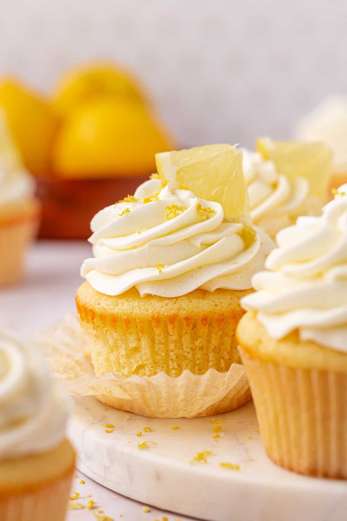 A close-up of lemon cupcakes with swirled white frosting, topped with lemon wedges and zest, sitting on a marble surface. In the background, whole lemons are slightly out of focus.