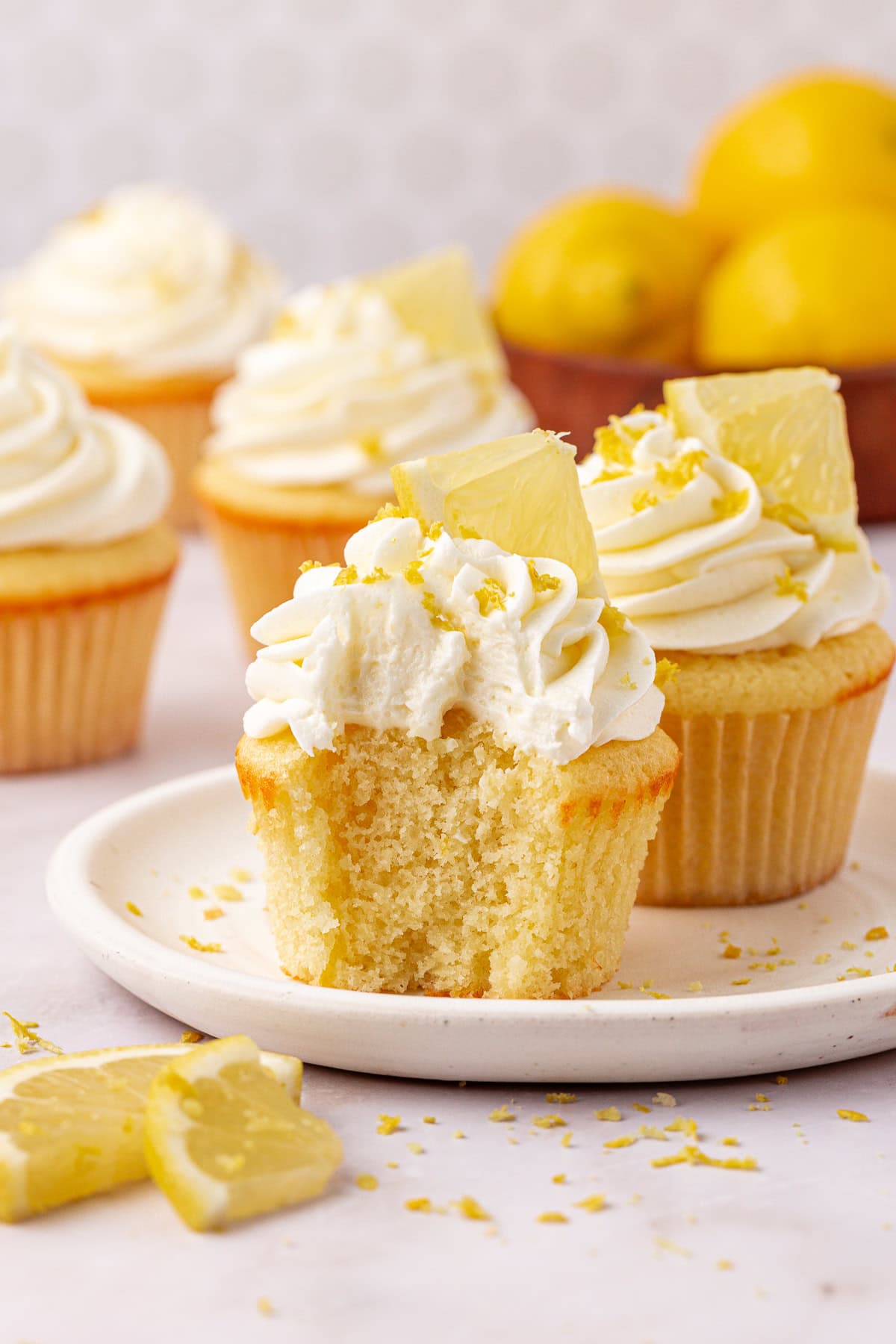 A plate with a lemon cupcake topped with whipped frosting and a lemon wedge; the cupcake has a bite taken out. More frosted cupcakes and a bowl of lemons are in the background. Lemon zest is sprinkled around.