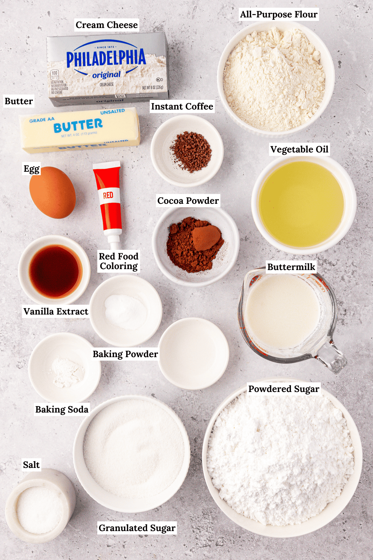 Top-down view of labeled baking ingredients in bowls and containers, including cream cheese, flour, butter, instant coffee, vegetable oil, egg, red food coloring, cocoa powder, buttermilk, vanilla, sugars, baking powder, baking soda, and salt.