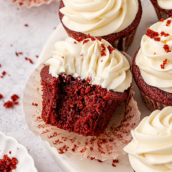 A group of red velvet cupcakes with swirls of cream cheese frosting. One red velvet cupcake has a big bite taken out, revealing its moist, rich interior. Red crumbs are scattered on the cupcakes and plate. Text reads Red Velvet Cupcakes.