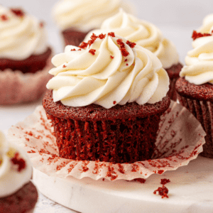A close-up of a red velvet cupcake with creamy white frosting and red crumbs on top, sitting on a parchment liner, with more cupcakes blurred in the background.