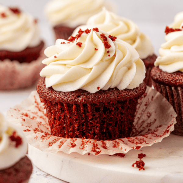 A close-up of a red velvet cupcake with creamy white frosting and red crumbs on top, sitting on a parchment liner, with more cupcakes blurred in the background.