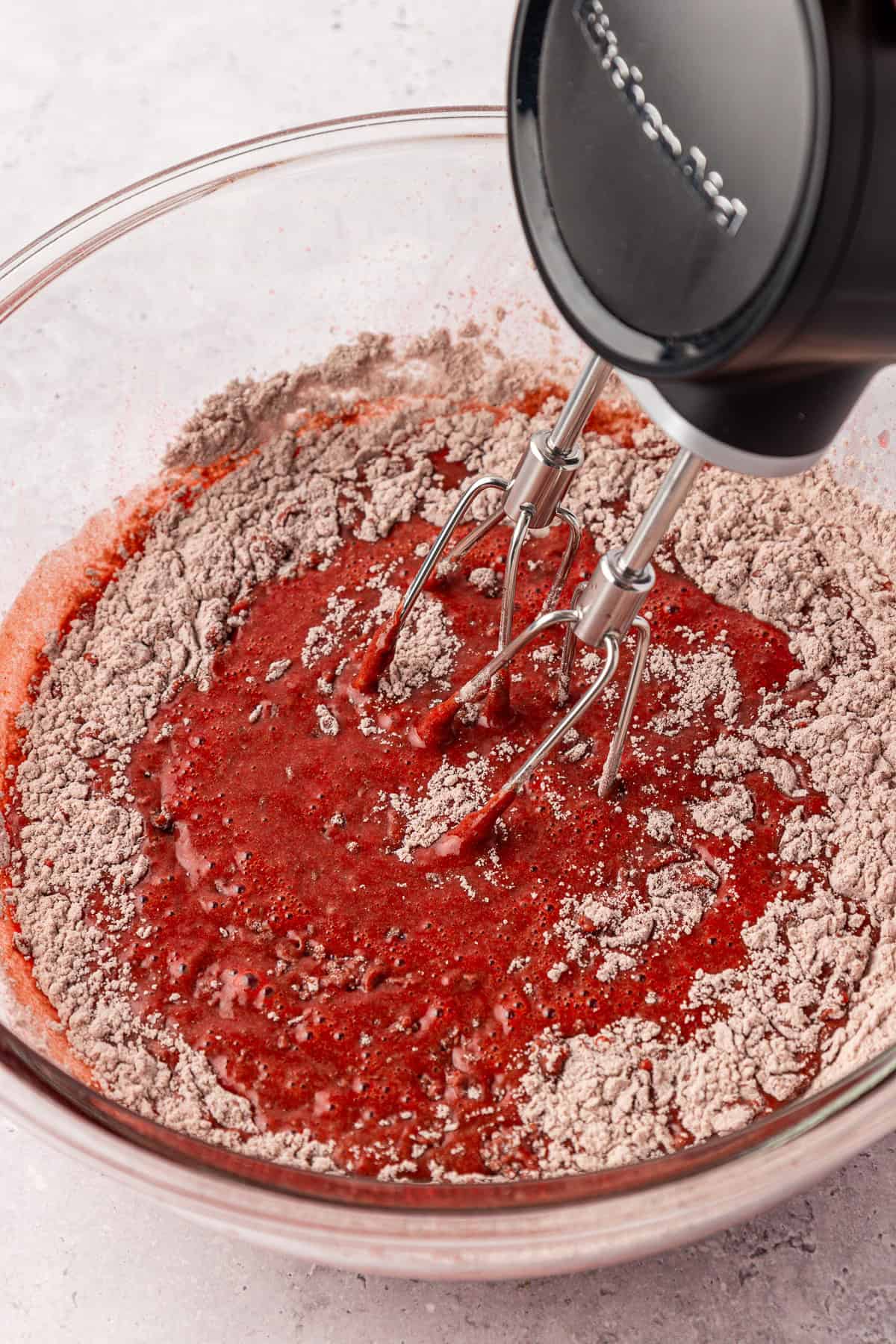 A hand mixer with beaters is mixing red liquid and chocolate cake mix in a clear glass bowl on a light countertop.