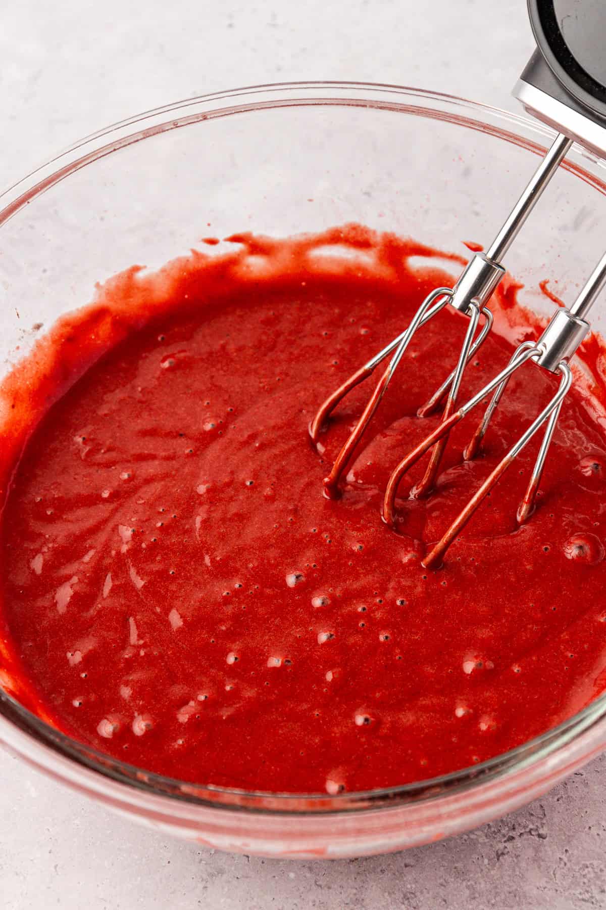 A glass bowl filled with bright red cake batter, with an electric hand mixer’s beaters resting inside the mixture on a light-colored countertop.
