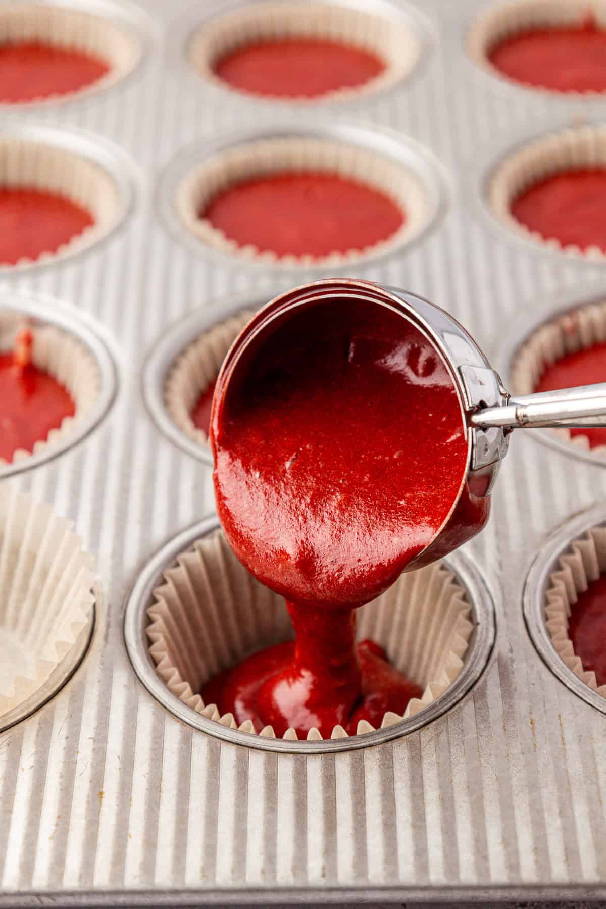 A metal scoop pours red cupcake batter into a paper-lined muffin tin. Other wells are already filled with the vibrant red batter, ready for baking.