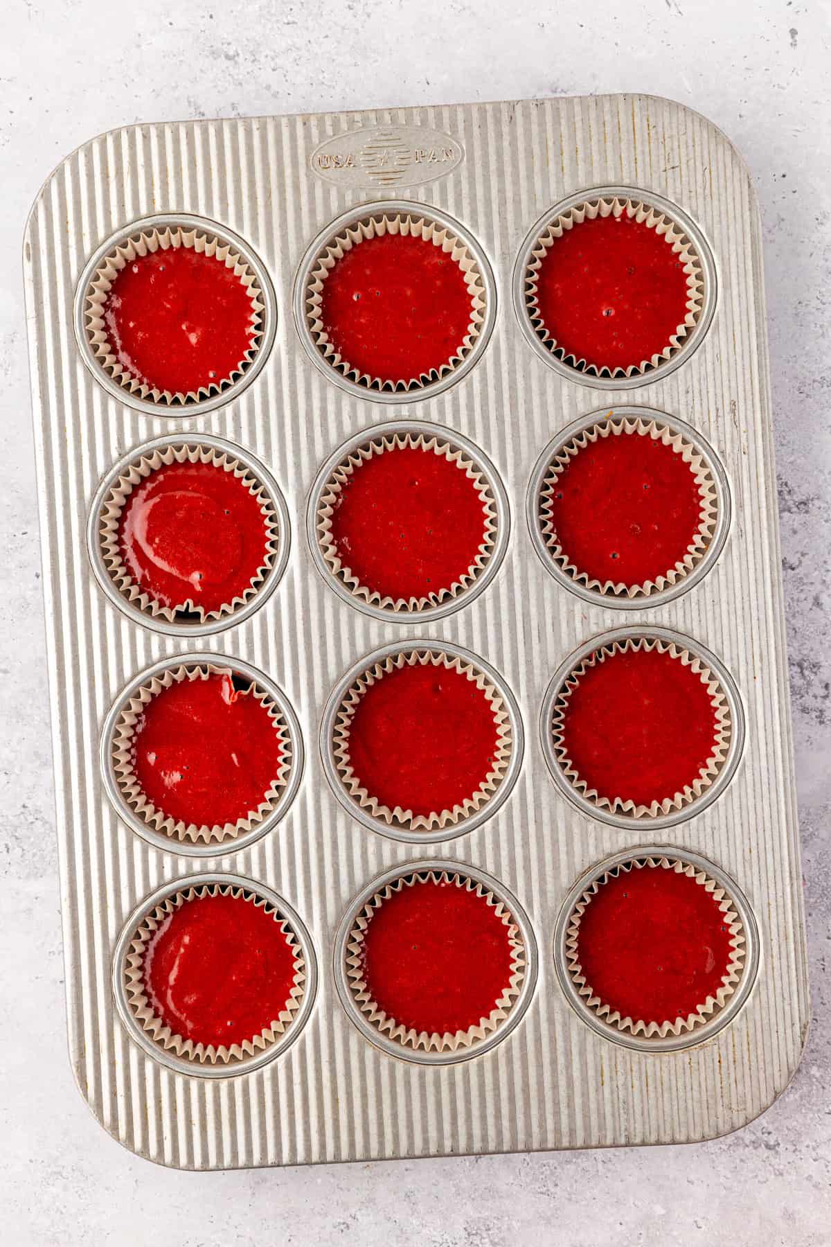 A metal muffin tin lined with 12 paper cupcake wrappers, each filled with red cake batter, is placed on a light-colored surface.