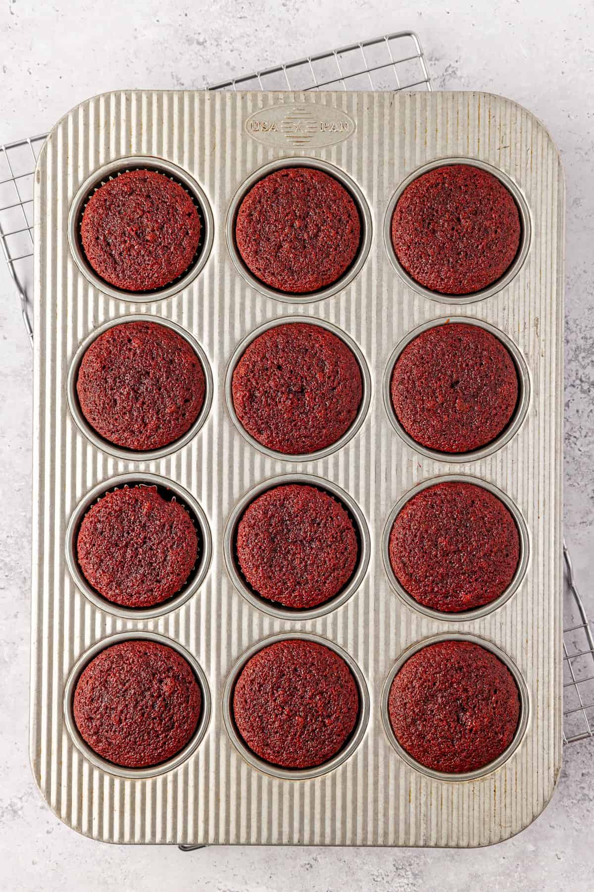 A top-down view of a muffin tray holding twelve freshly baked red velvet cupcakes, resting on a wire cooling rack on a light gray surface.
