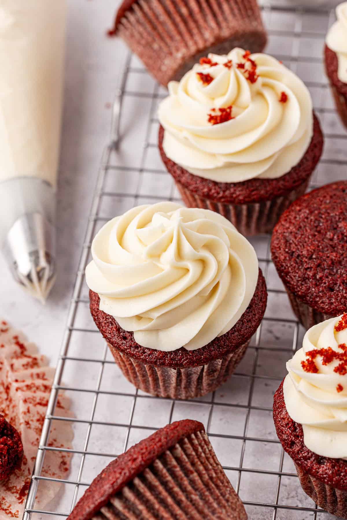 Red velvet cupcakes with swirls of white frosting on a cooling rack, with some cupcake wrappers and a piping bag nearby.