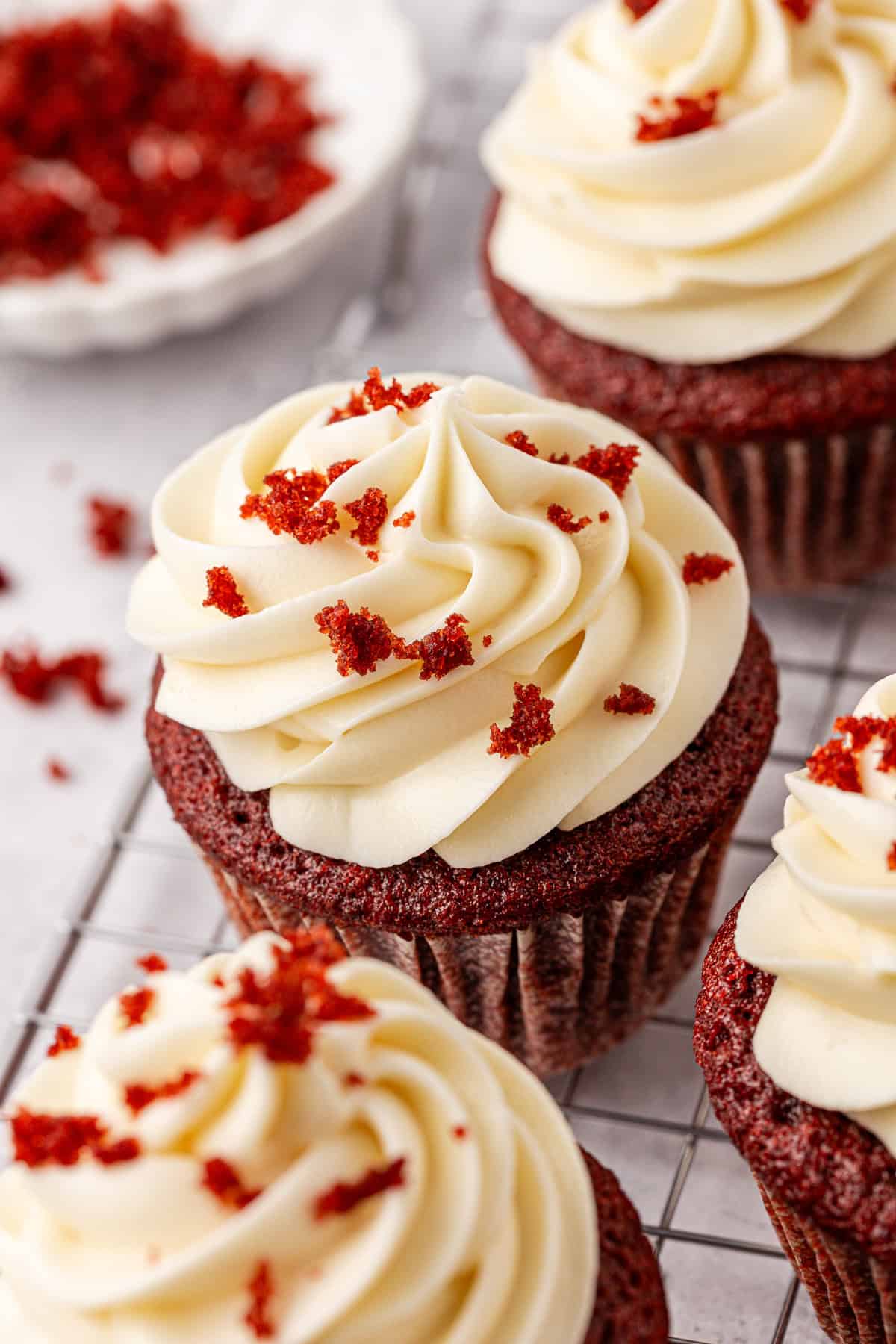 Close-up of red velvet cupcakes topped with swirls of creamy white frosting and sprinkled with red velvet cake crumbs, displayed on a wire cooling rack. A dish with more crumbs is visible in the background.