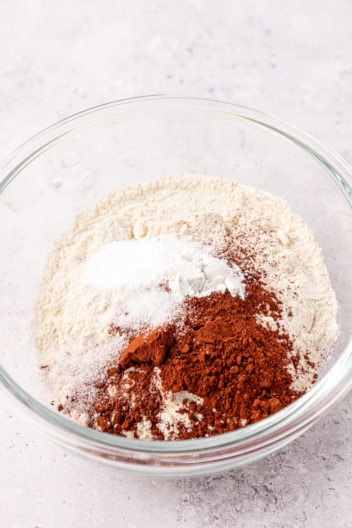 A glass bowl containing dry baking ingredients, including flour, cocoa powder, baking powder, baking soda, and salt, on a light-colored countertop.