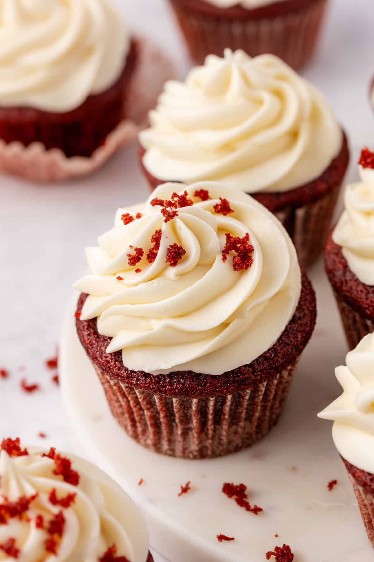 A close-up of red velvet cupcakes topped with swirls of creamy white frosting and sprinkled with red velvet crumbs, displayed on a white surface.
