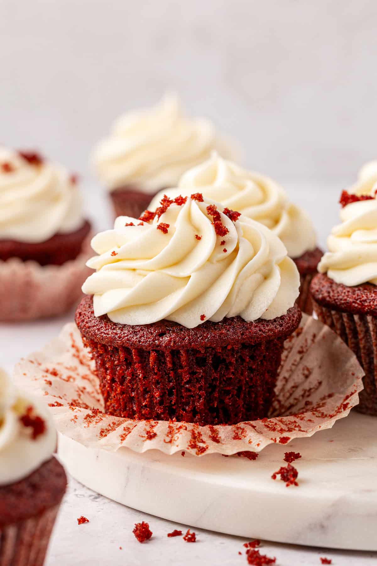 Red velvet cupcakes topped with swirls of creamy white frosting, some with red cake crumbs sprinkled on top, are arranged on a light surface with a cupcake liner partially peeled back in the foreground.