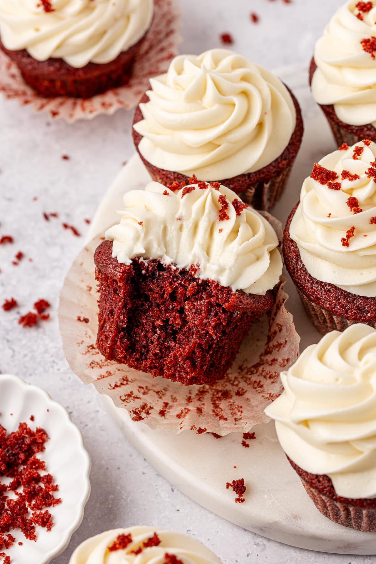 Red velvet cupcakes with swirled cream cheese frosting are arranged on a white surface. One cupcake in the center has a bite taken out, showing its moist, red crumb. Red velvet crumbs are scattered around.