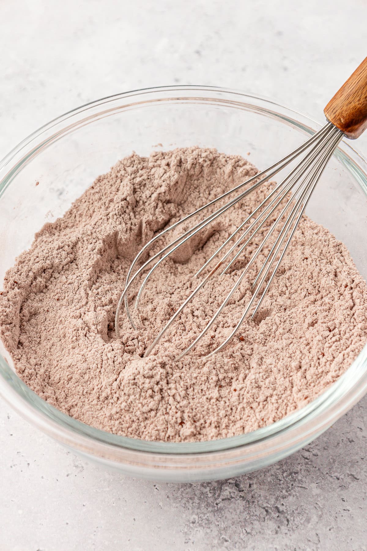 A glass bowl filled with light brown powder, being mixed with a metal whisk with a wooden handle, sits on a light gray countertop.