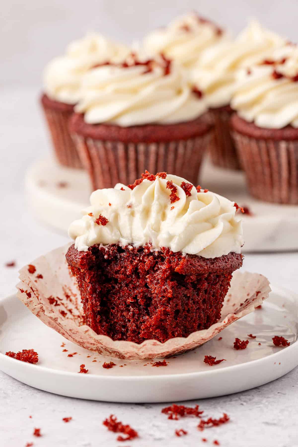 A red velvet cupcake with white frosting sits on a plate, partially unwrapped and bitten into, with crumbs scattered around. Three more frosted cupcakes are blurred in the background.