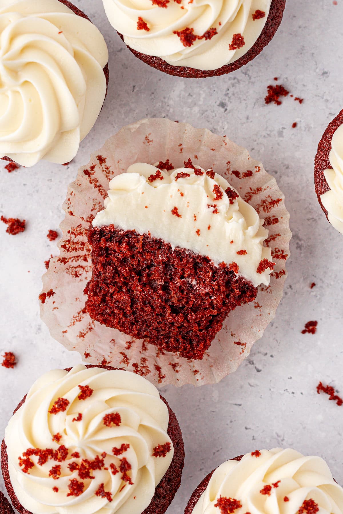 A red velvet cupcake with cream cheese frosting sits in a wrapper, partially eaten to show its moist crumb. Whole cupcakes with swirled frosting and red velvet crumbs surround it on a light surface.