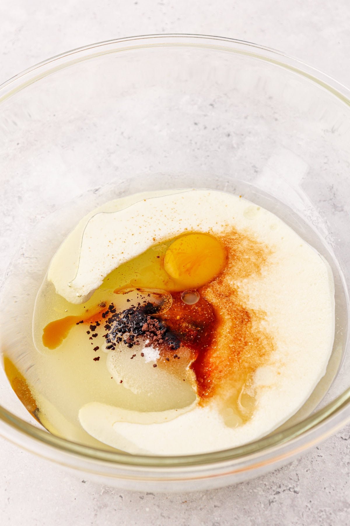 A clear glass bowl contains an egg, sugar, vanilla extract, and other baking ingredients on a light-colored surface, ready to be mixed.