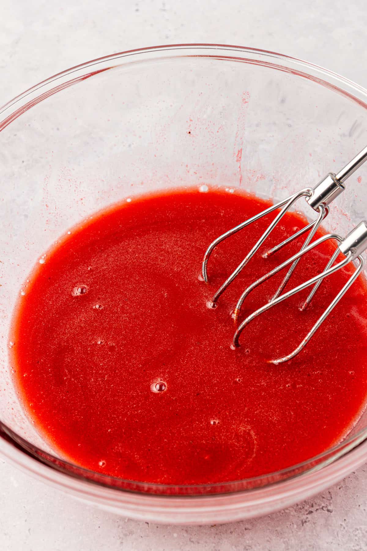 A glass bowl filled with bright red liquid being mixed by a metal hand mixer, set on a light-colored surface.