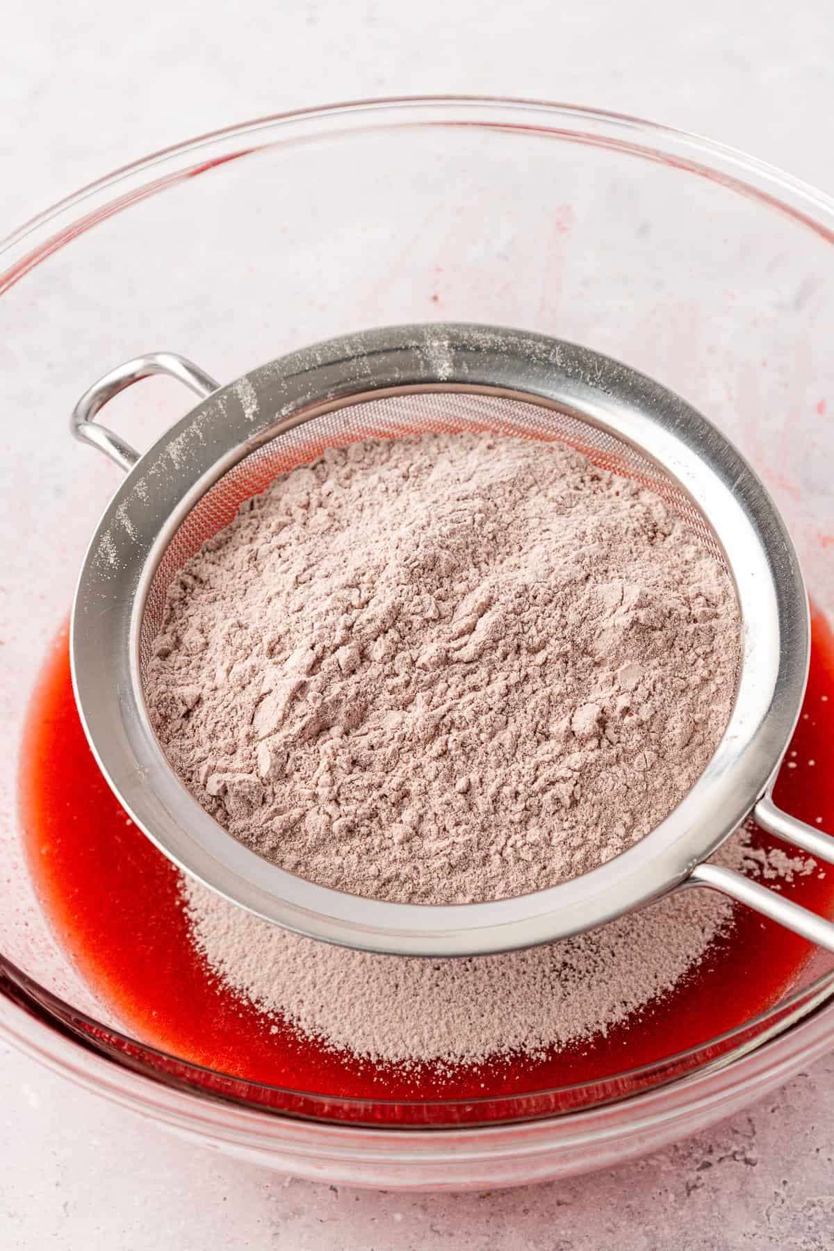 A metal sifter filled with cocoa powder rests over a glass bowl containing red liquid, likely for a baking recipe, on a light-colored surface.