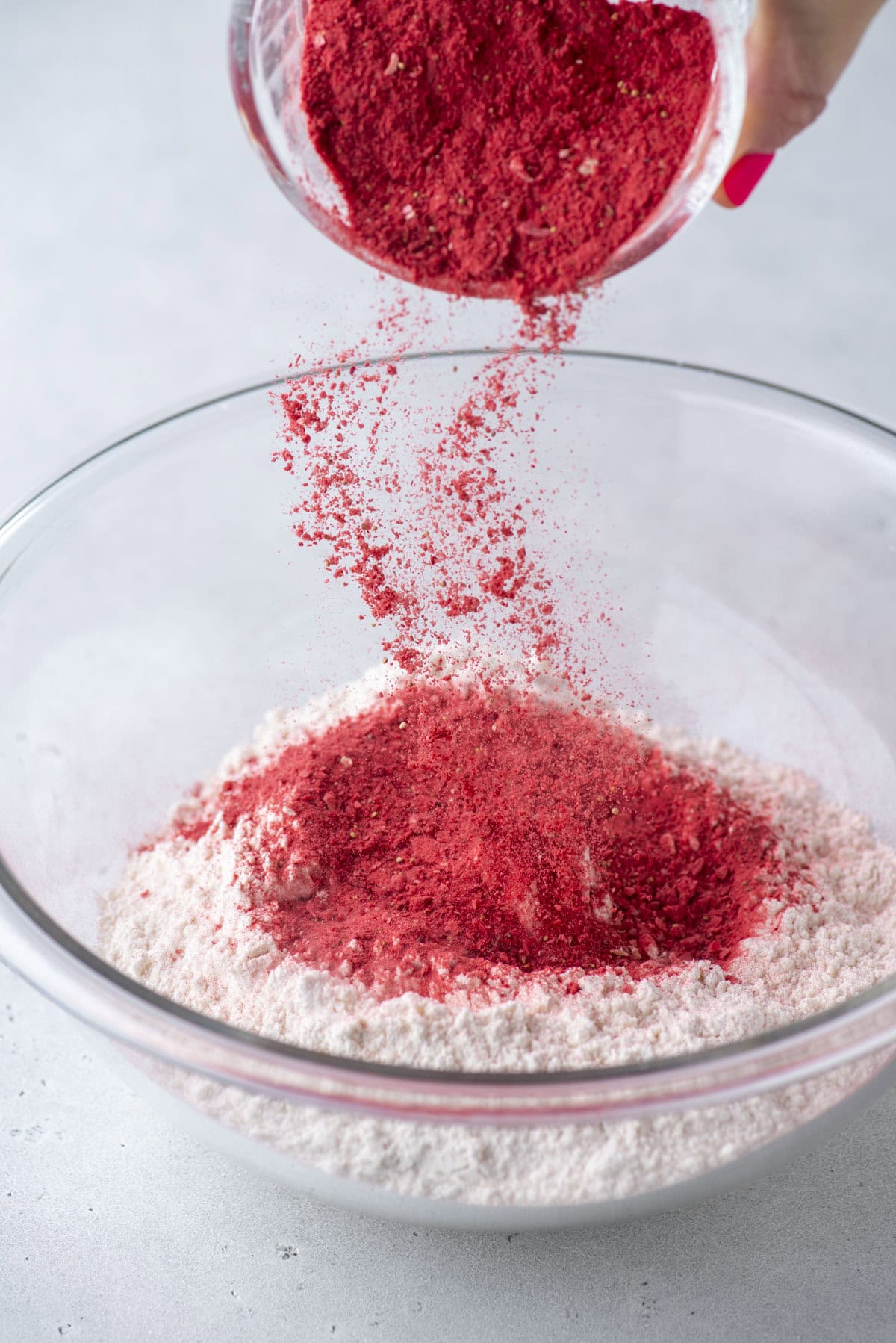 A hand pours vibrant red powder into a clear glass bowl filled with flour, creating a colorful dusting above the mixture on a light gray surface.