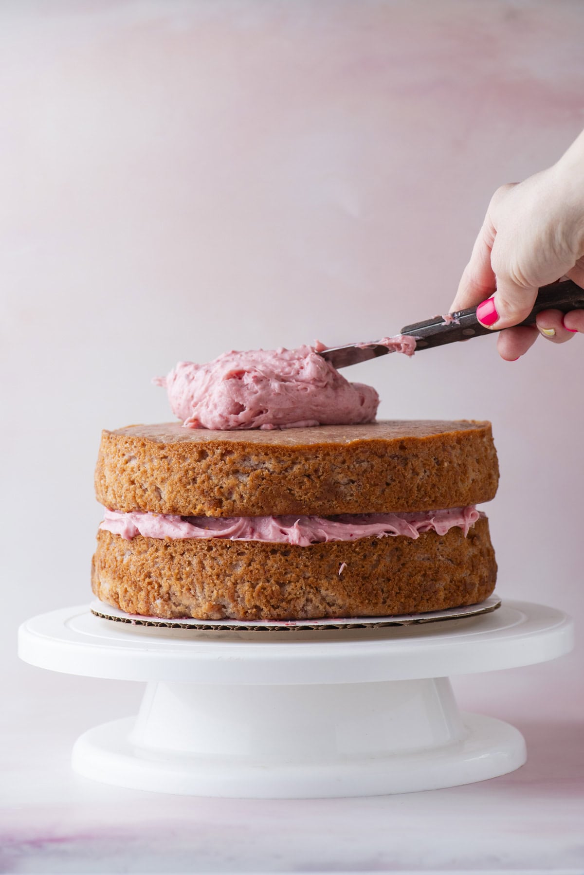 A hand spreads pink frosting on top of a two-layer cake with pink filling, placed on a white cake stand against a soft pink background.