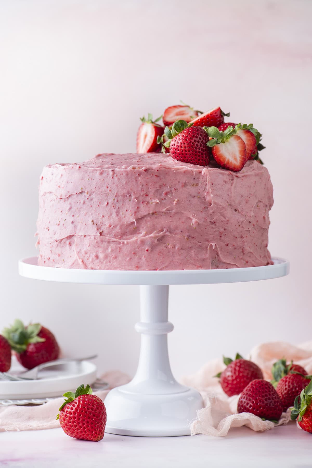 A round cake with pink strawberry frosting sits on a white cake stand, topped with fresh strawberries. Whole and halved strawberries are scattered around the base, with a soft pink background and plates in the distance.