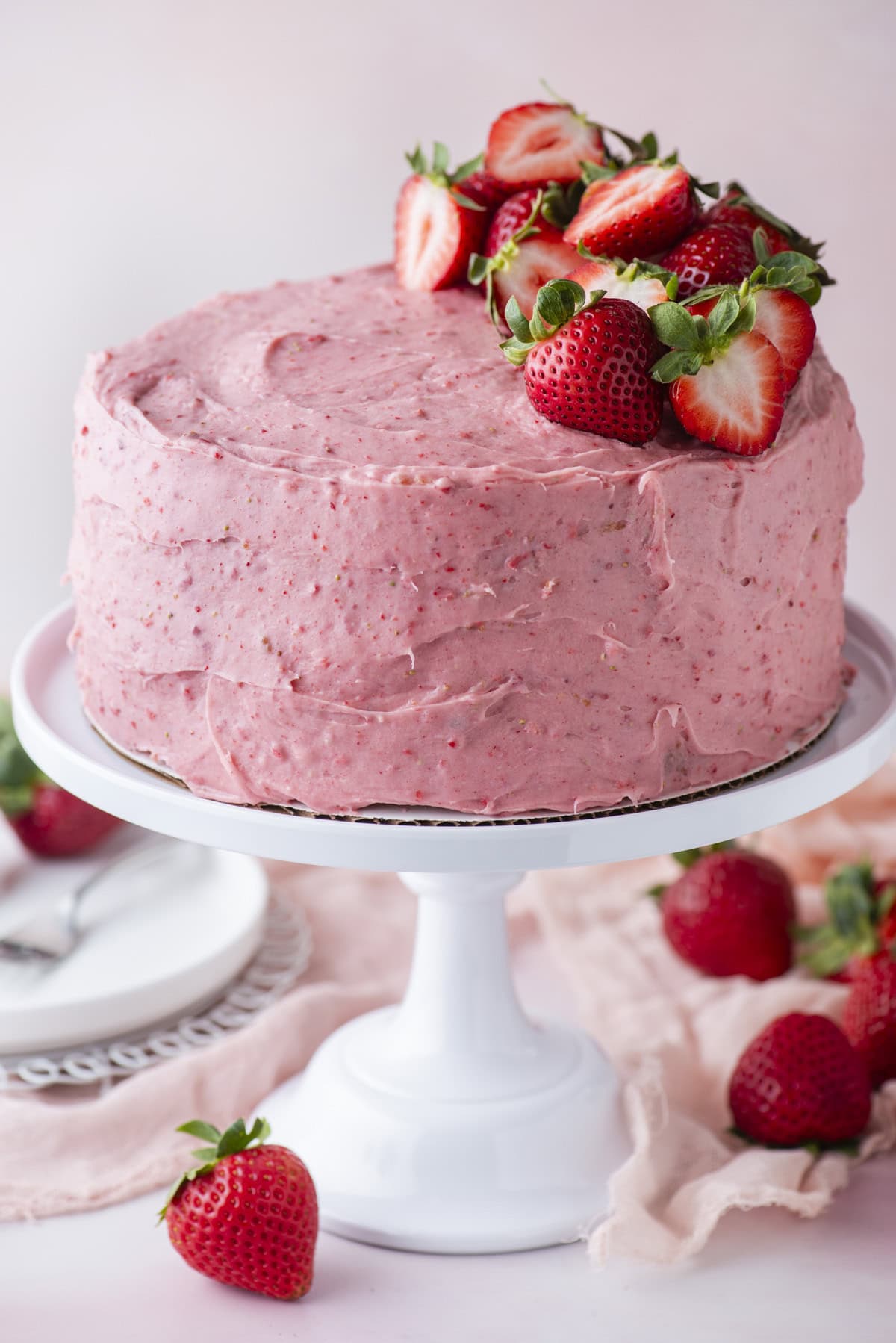 A round cake with pink strawberry frosting sits on a white cake stand, topped with fresh strawberries. Whole strawberries are scattered around the base, and plates and utensils are visible in the background.