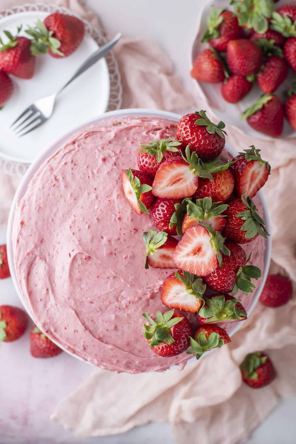 A cake topped with pink strawberry frosting and fresh strawberries, some halved, sits on a light pink surface with whole strawberries and a plate with a fork nearby.