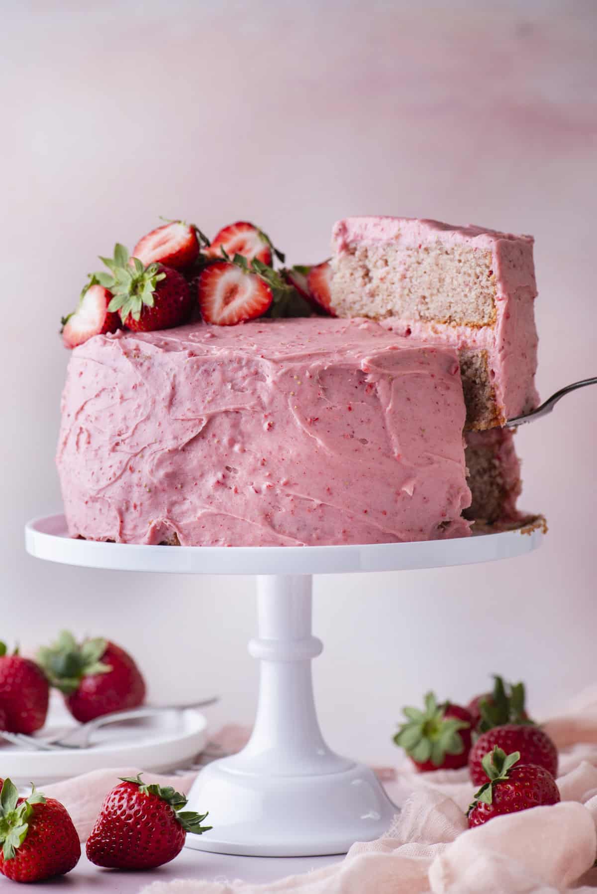 A strawberry cake with pink frosting and fresh strawberries on top sits on a white cake stand. A slice is being lifted out, revealing the cake layers inside. Strawberries are scattered around the base.