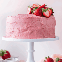 A strawberry cake with pink strawberry frosting, topped with fresh strawberries, sits on a white cake stand. Extra strawberries are scattered around the stand. Text above reads “Strawberry Cake.”.