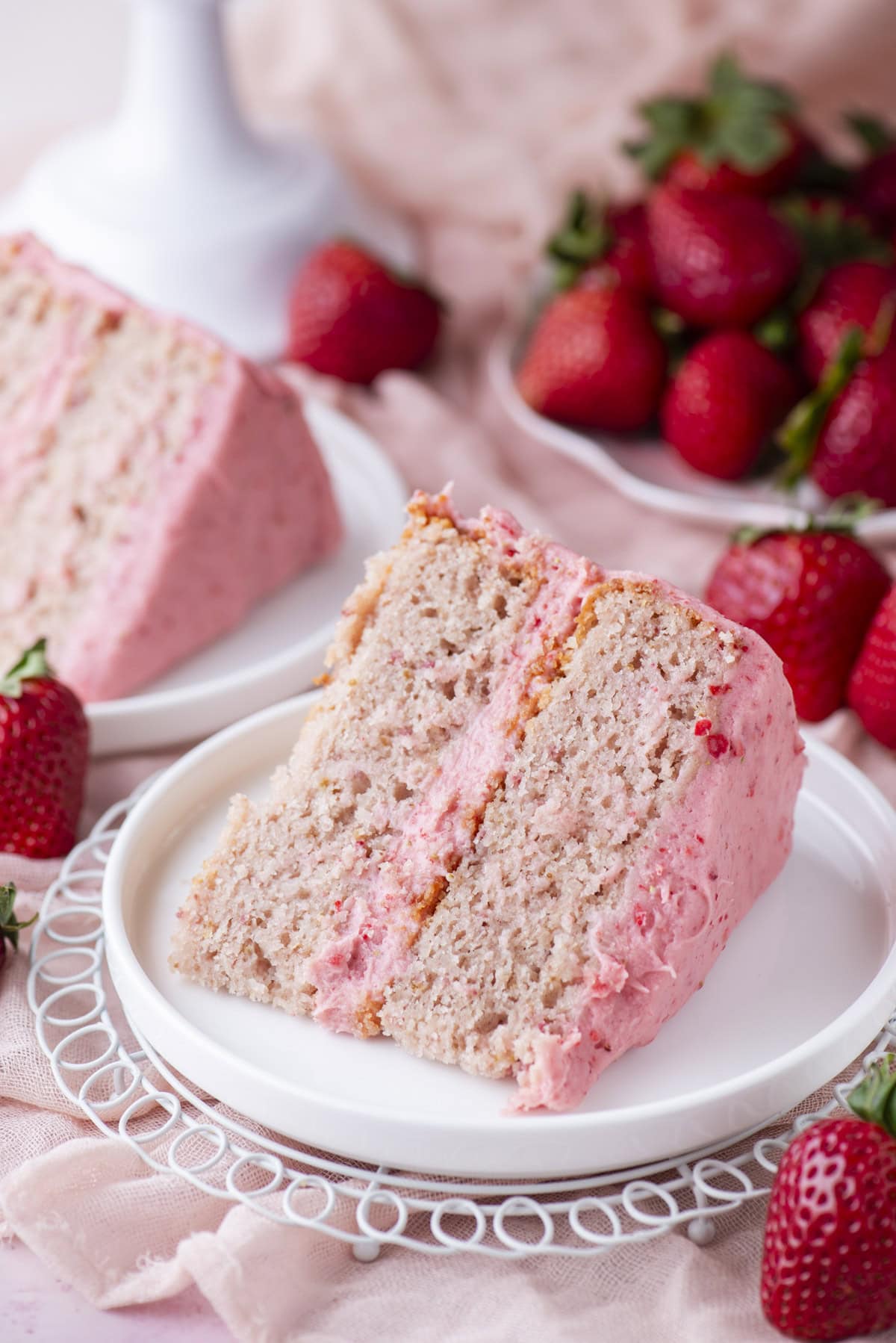 A slice of strawberry cake with pink frosting sits on a white plate, with fresh strawberries scattered around and a second slice of cake in the background.