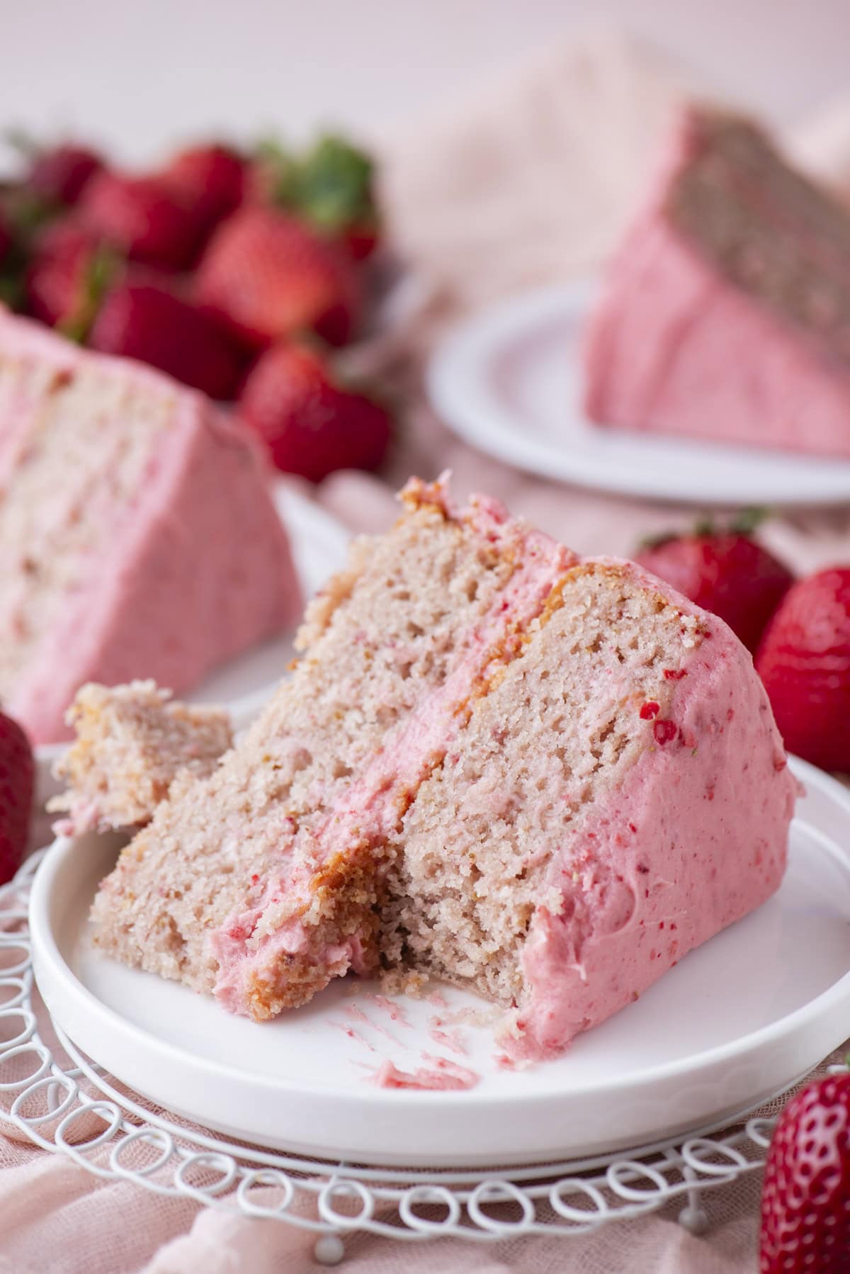 A slice of strawberry cake with pink frosting sits on a white plate, surrounded by fresh strawberries, with more cake slices in the blurred background.